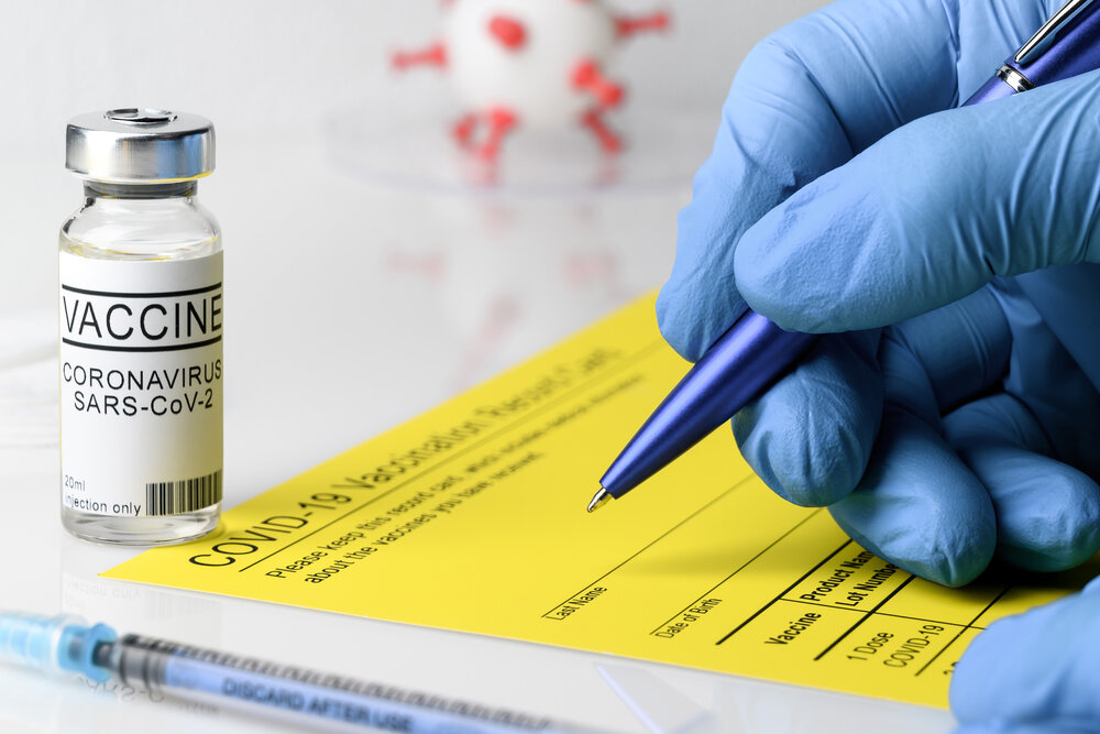 Coronavirus vaccination theme. Doctor's hands in medical gloves filling out the vaccination record card, a vial with covid-19 vaccine and syringe on a white table, close up